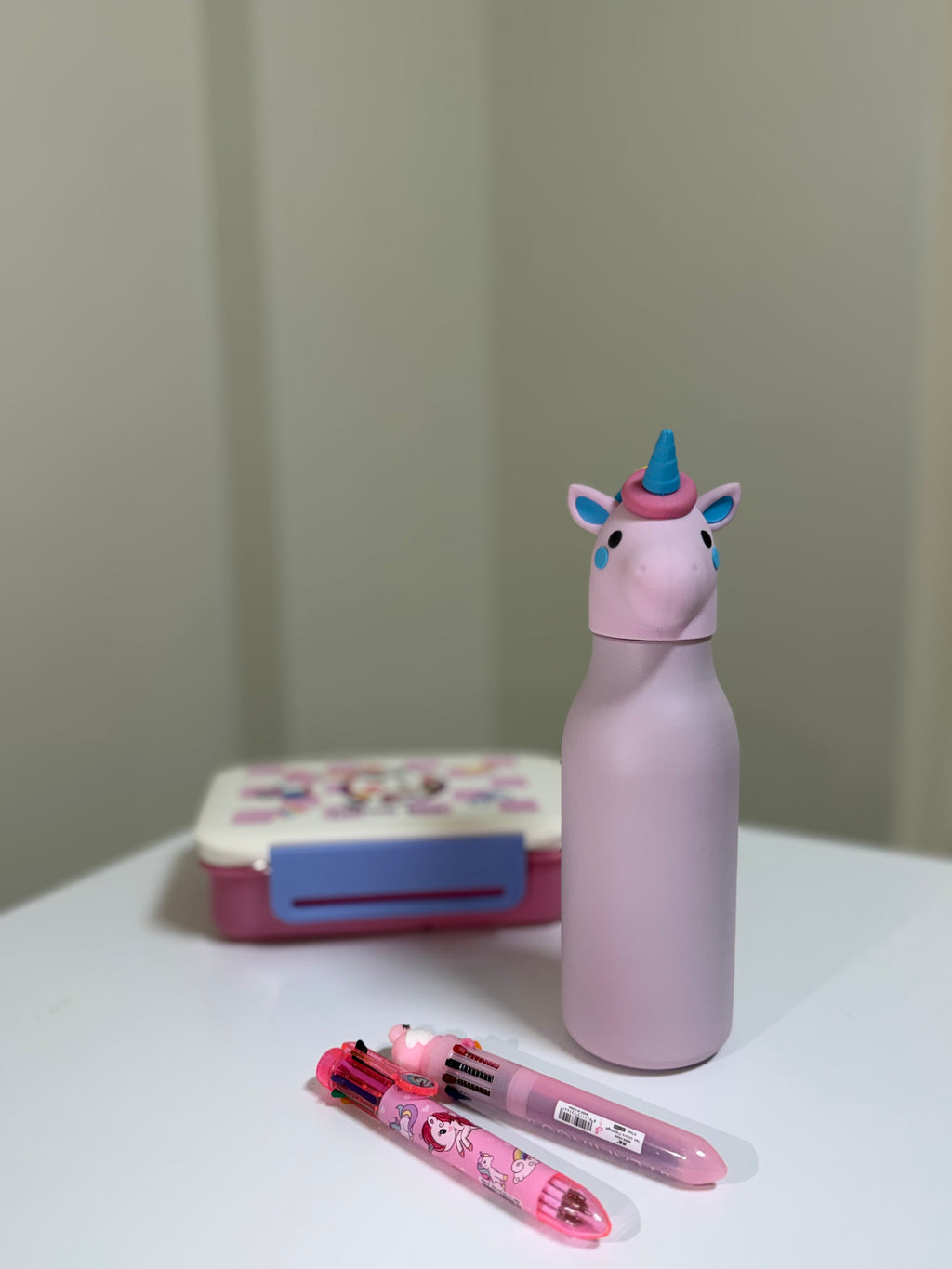 A pink unicorn-shaped bottle with a blue horn stands on a white table next to a pink and blue lunchbox and two pink cartoon pens.