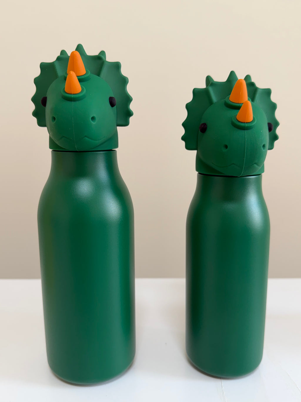 Two green dinosaur-shaped water bottles with orange horns and spiky backs, standing on a white surface against a light beige background.