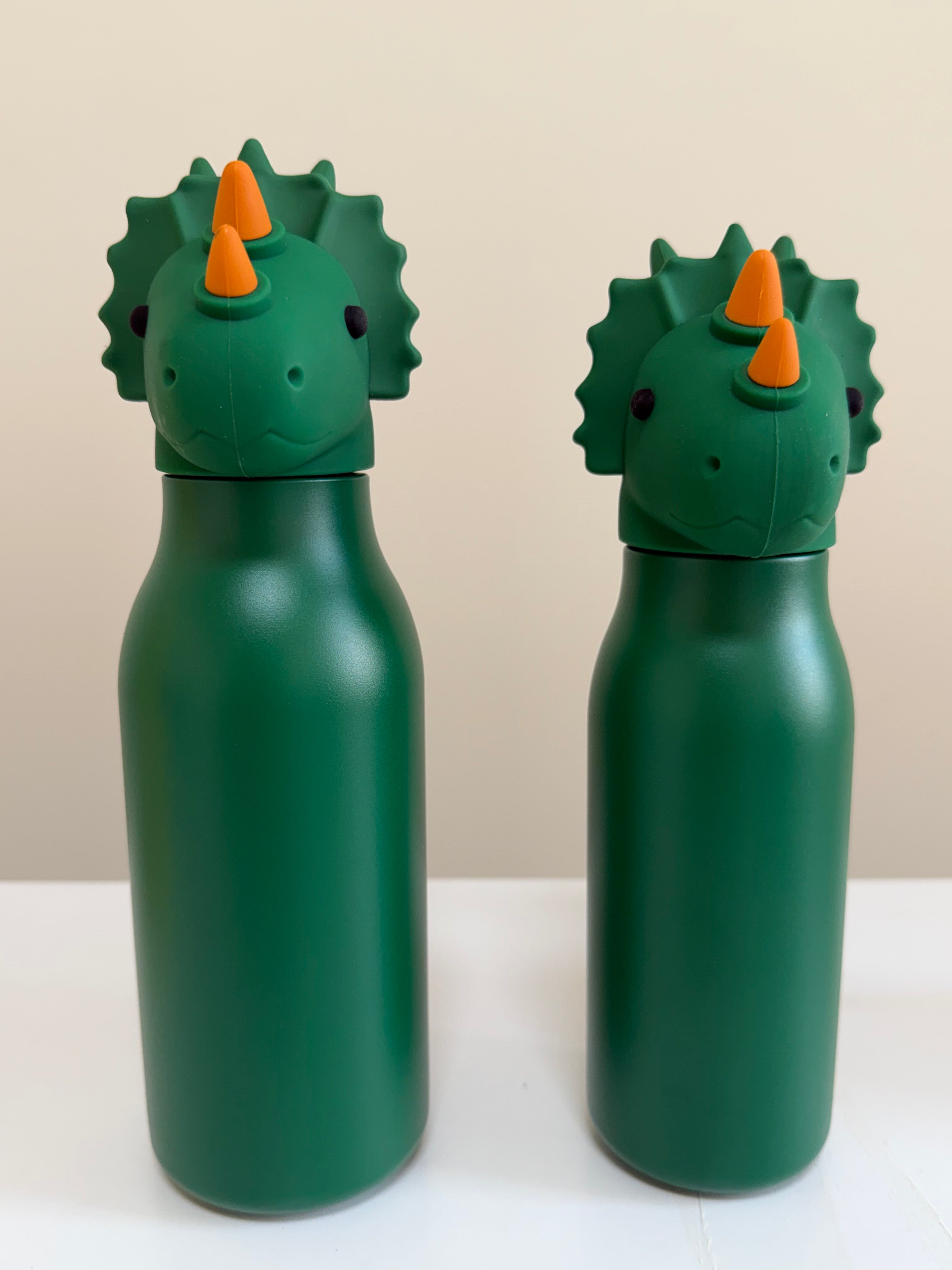 Two green dinosaur-shaped water bottles with orange horns and spiky backs, standing on a white surface against a light beige background.
