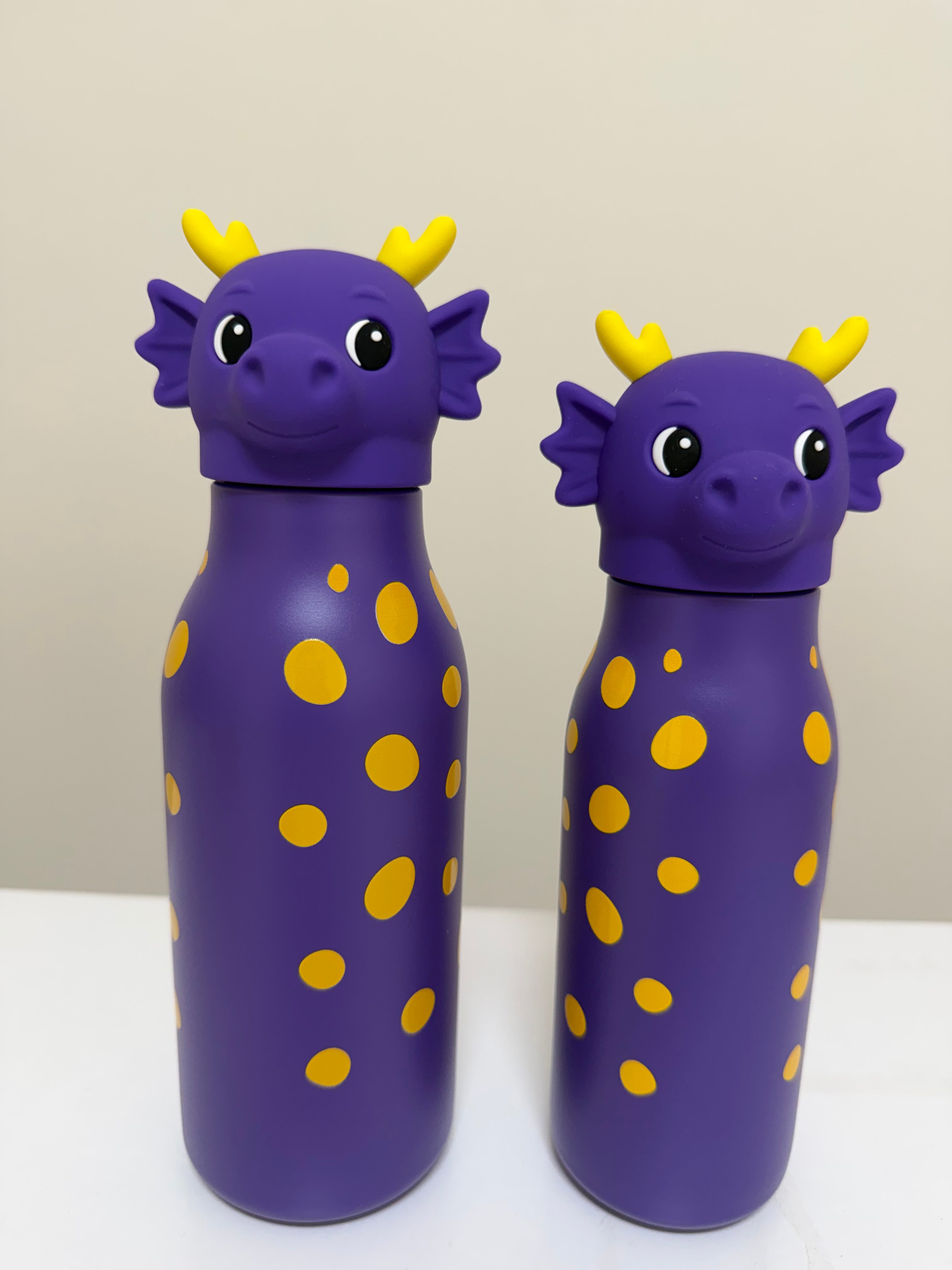 Two purple dragon-shaped water bottles with yellow spots and yellow horns, standing on a white surface against a light beige background.