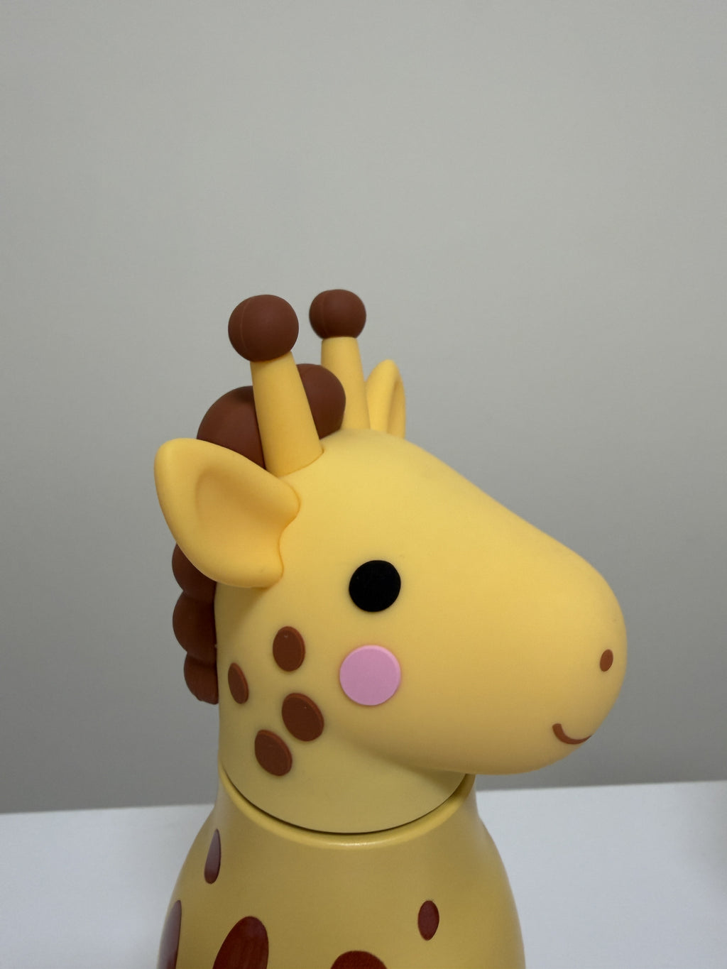A yellow giraffe-shaped container or toy with brown spots, a pink cheek, and a black eye, shown in a close-up side view against a plain light gray background.